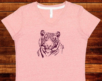 May include: A light pink V-neck t-shirt featuring a detailed purple illustration of a tiger's face. The shirt has short sleeves and a white neckline. The design is centered on the front of the shirt.