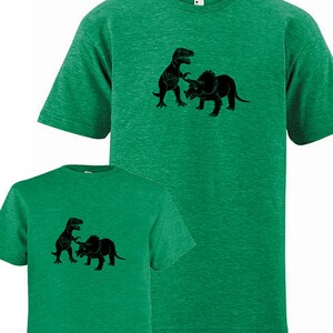 Puede incluir: Dos camisetas verdes con una silueta negra de un Tyrannosaurus Rex luchando contra un Triceratops.