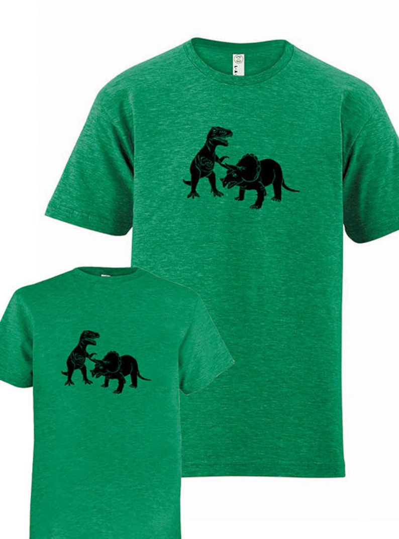 Puede incluir: Dos camisetas verdes con una silueta negra de un Tyrannosaurus Rex luchando contra un Triceratops.