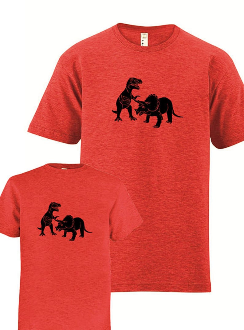 Puede incluir: Dos camisetas rojas con una silueta negra de un dinosaurio atacando a un triceratops. Las camisetas son un conjunto a juego para adultos y ni&ntilde;os.