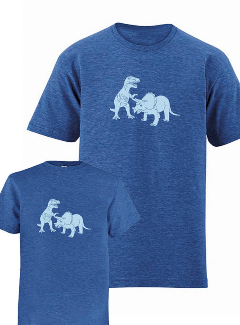 Puede incluir: Dos camisetas azules con un gr&aacute;fico de un dinosaurio luchando contra un triceratops. El gr&aacute;fico es de color azul claro.
