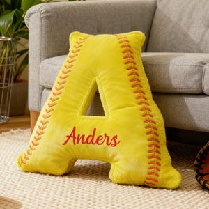 Könnte beinhalten: Gelbes Kissen in Form des Buchstabens "A", im Softball-Stil. Es hat rote Nähte und den Namen "Anders" in roter Schrift. Das Kissen liegt auf einem hellen Teppich.