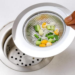 May include: A stainless steel sink drain with a removable strainer basket. The strainer contains chopped vegetables, including green celery stalks and yellow sweetcorn kernels. The drain is partially open, revealing the drain opening.