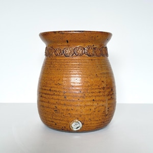 May include: A brown ceramic vase with a textured finish and a floral pattern around the neck. The vase has a rounded shape and a flared rim. The base features a small, circular marking. The vase is likely handmade.