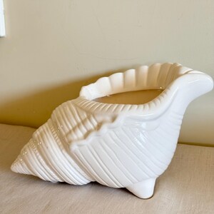 Peut inclure: Un cache-pot en céramique blanche en forme de coquillage conque. L'objet présente une finition brillante, un extérieur texturé avec des crêtes en relief, une large ouverture sur le dessus et de petits pieds à la base pour assurer la stabilité.