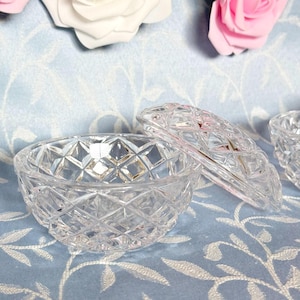 May include: A clear, cut-glass trinket dish with a matching lid, set against a light blue background. The dish and lid feature a diamond pattern. Decorative pink and white roses are in the background.