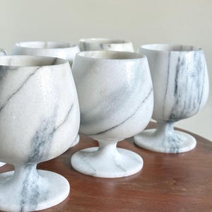May include: A collection of white marble goblets featuring natural grey veining throughout the stone. Each goblet has a rounded bowl, a short stem, and a circular base, showcasing the unique polished texture and patterns of the natural stone material.