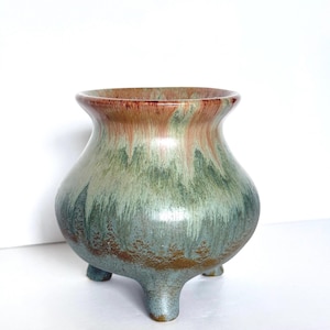 May include: A ceramic vase with a rounded body and three small feet. The vase has a wide opening and a flared rim. The colours include shades of green, blue, and brown, with a gradient effect. The vase is likely handmade and suitable for home decor.