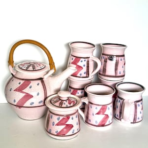 May include: A pink ceramic tea set with a teapot, sugar bowl, and mugs. The set features a zigzag design in red and blue dots. The teapot has a bamboo handle.