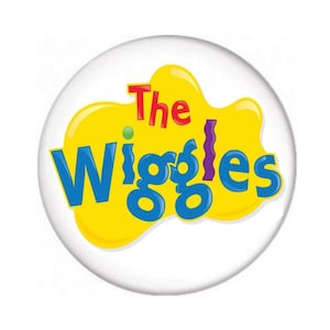 May include: A circular pinback button featuring a white background with a central yellow blob shape. The text The Wiggles is printed in a stylized blue font with the word The in red, centered on the yellow shape against the white circular surface.