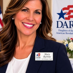 May include: A rectangular name badge featuring the DAR logo with a blue star and red stripes, the name Mary Barrow, and the text Lady Washington Chapter. The badge is white with a black border and is pinned to a dark navy blue blazer.