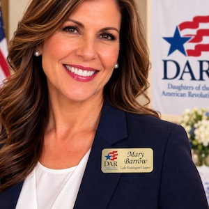 May include: A rectangular gold-toned name badge featuring the DAR logo with a blue star and red stripes, the name Mary Barrow, and the text Lady Washington Chapter. The badge is pinned to a dark navy blue blazer worn over a white shirt.
