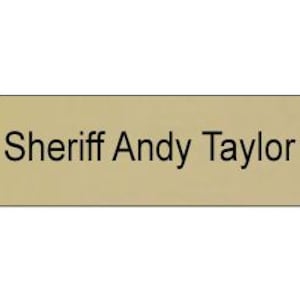 May include: A rectangular name badge with a light tan background and rounded corners. The text "Sheriff Andy Taylor" is printed in a clear, black, sans-serif font centred on the face of the badge. The design is simple and functional.