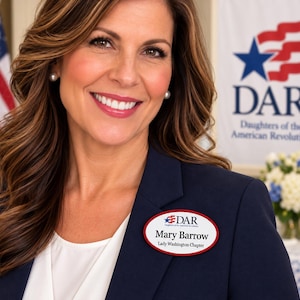 May include: An oval-shaped name badge with a red border, featuring the DAR logo, the name Mary Barrow, and the text Lady Washington Chapter. The badge is pinned to a dark navy blue blazer worn over a white top, with a flag and flowers visible.