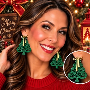 May include: A pair of green Christmas tree-shaped earrings featuring a gold-colored bow at the top. The earrings have a detailed, swirling cutout design and a glittery texture. A circular inset shows a clear view of one earring on an earlobe.