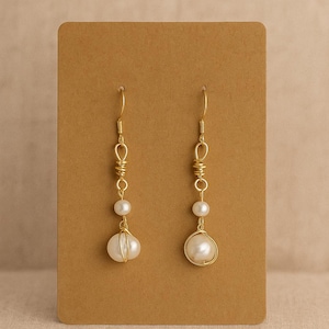 May include: A pair of gold-colored wire earrings with white pearls. Each earring has a long, thin hook, with a series of small pearls and wire detailing. The bottom pearl is larger and encased in a gold wire cage.