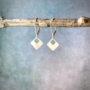 May include: A pair of silver dangle earrings with a square shape and a cut-out design. The earrings are hanging from a branch.