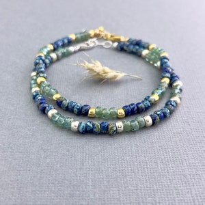 May include: Two beaded bracelets with blue, green, and gold beads. The bracelets are made of small, round beads in shades of blue and green, interspersed with gold and silver-colored beads. A sprig of dried grass rests between the bracelets.
