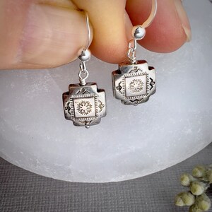 May include: A pair of silver dangle earrings with a square design featuring a detailed floral pattern. The earrings have a simple hook closure.