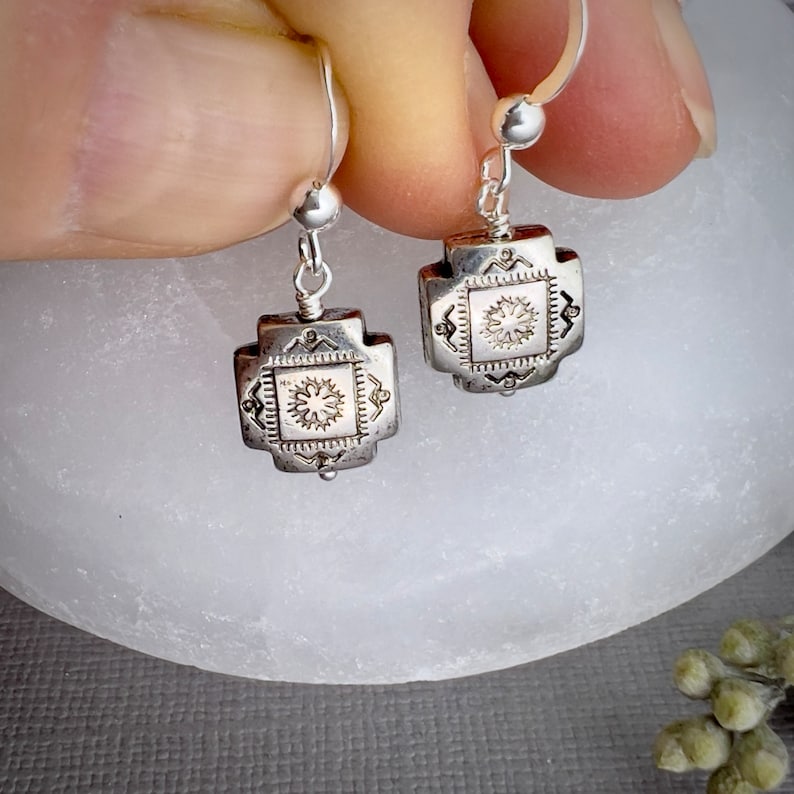 May include: A pair of silver dangle earrings with a square design featuring a detailed floral pattern. The earrings have a simple hook closure.