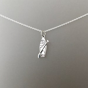 May include: A silver-colored pendant necklace featuring a stand-up paddleboard and paddle charm. The charm has a detailed design with a spiral and a paddle. The necklace is on a silver chain, perfect for water sports enthusiasts.