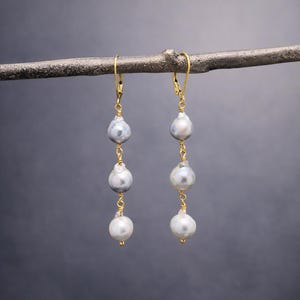 May include: Elegant gold-tone earrings with three pear-shaped, gray-white pearls suspended from a gold hook. The pearls are connected by delicate gold wire, creating a sophisticated and timeless design.