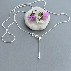 May include: A delicate silver necklace with a slender chain and adjustable length. The necklace features a small, round pendant at the end of the adjustable portion. A small stone and sprig of purple and white flowers rest on a gray stone in the background.