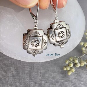 May include: Silver dangle earrings with a square design featuring a detailed floral pattern. The earrings have a hook closure.