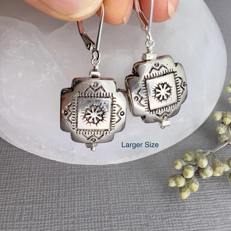 May include: Silver dangle earrings with a square design featuring a detailed floral pattern. The earrings have a hook closure.