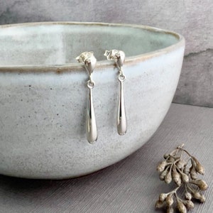 May include: A pair of silver teardrop earrings with a smooth, polished finish. The earrings are hanging from a ceramic bowl.