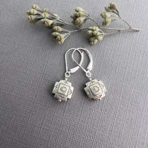 May include: A pair of silver earrings with a square design featuring a detailed geometric pattern. The earrings have a leverback closure.
