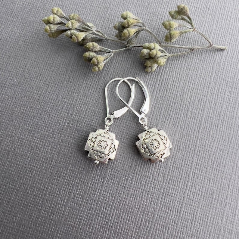 May include: A pair of silver earrings with a square design featuring a detailed geometric pattern. The earrings have a leverback closure.