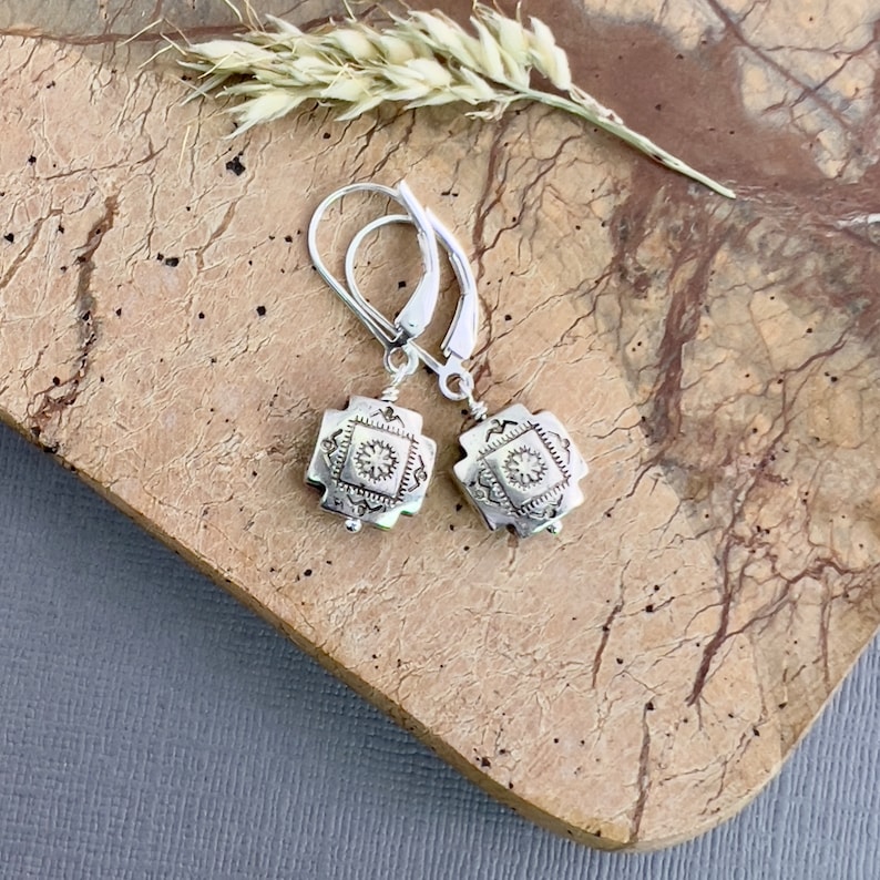 May include: Silver dangle earrings with a square-shaped charm featuring a detailed floral design. The earrings have a leverback closure.