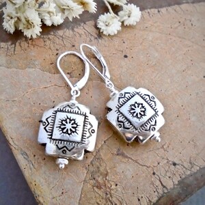 May include: A pair of silver earrings with a geometric design. Each earring features a square-shaped pendant with a raised, intricate pattern of a starburst within a square.