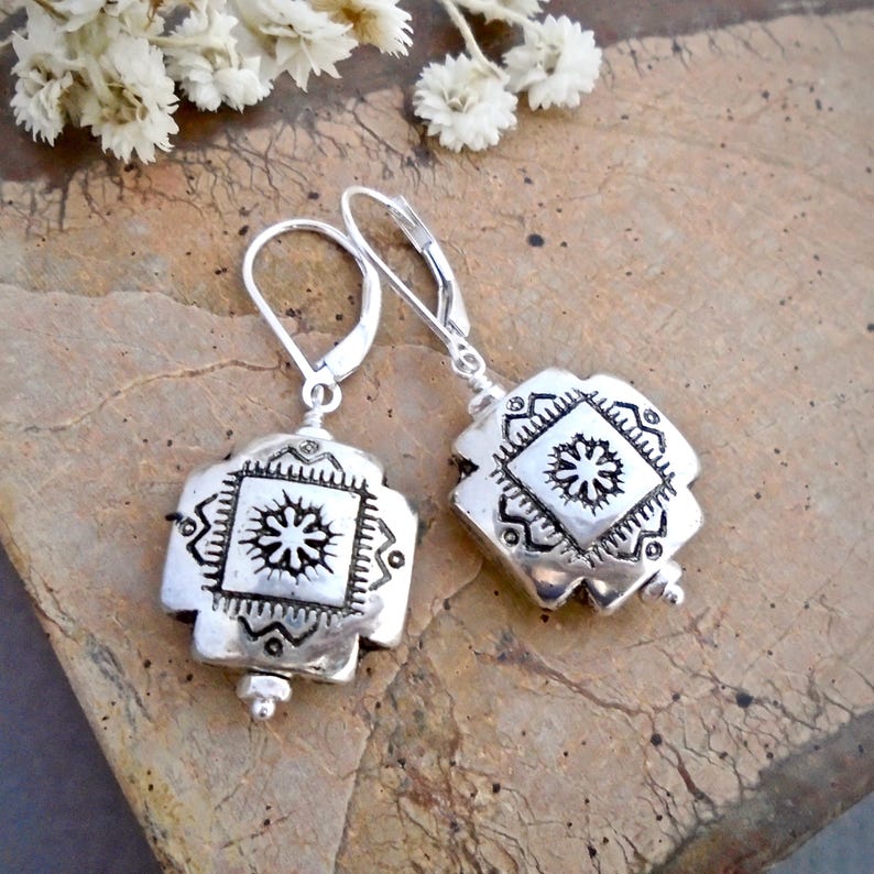 May include: A pair of silver earrings with a geometric design. Each earring features a square-shaped pendant with a raised, intricate pattern of a starburst within a square.