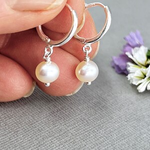 May include: A pair of silver hoop earrings with a dangling, round, off-white pearl. The earrings are held up against a gray background, with a few purple and white flowers in the background.