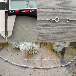 May include: A close-up of a silver satellite chain necklace with small round beads. The chain is 3.56 mm wide. The necklace is shown on a textured surface with white flowers.