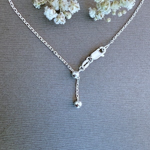May include: A delicate silver necklace with a small dangling ball pendant. The necklace features a fine chain and a lobster clasp closure. The background is a muted gray with sprigs of white flowers.
