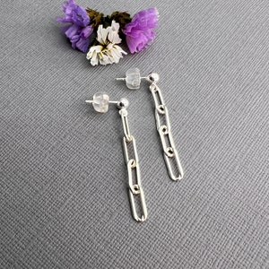 May include: A pair of silver chain link earrings with post backs. The earrings are displayed on a gray surface with a small arrangement of purple and white flowers in the background. The earrings have a polished finish.