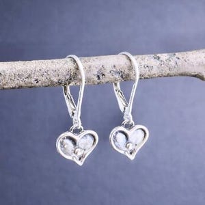 Puede incluir: Pendientes sencillos de plata de ley con forma de coraz&oacute;n. Los pendientes se muestran en una rama de color claro sobre un fondo azul oscuro. Los pendientes tienen un cierre de palanca y un peque&ntilde;o dise&ntilde;o de coraz&oacute;n texturizado.