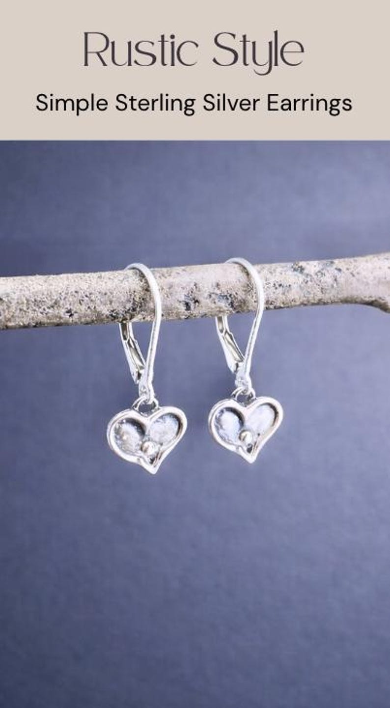 Puede incluir: Pendientes sencillos de plata de ley con forma de coraz&oacute;n. Los pendientes se muestran en una rama de color claro sobre un fondo azul oscuro. Los pendientes tienen un cierre de palanca y un peque&ntilde;o dise&ntilde;o de coraz&oacute;n texturizado.