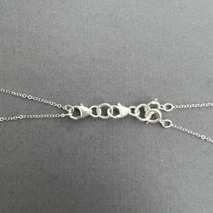 May include: Close-up of silver-colored necklace clasps and chain links against a gray background. The clasps are connected with small circular links. The chains extend from the clasps, suggesting a jewelry design.