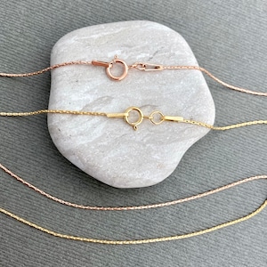 May include: Three gold chains of different lengths, two are yellow gold and one is rose gold. The chains are laying on a gray stone.