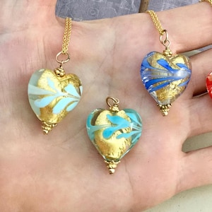 May include: Three heart-shaped glass pendants suspended on gold-toned chains. Each pendant features a clear glass exterior with internal gold leaf foil accents and swirling blue or light blue patterns embedded within the glass structure.