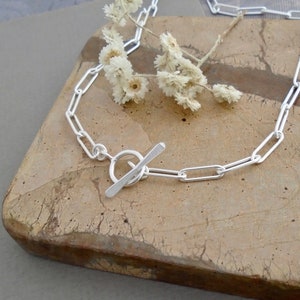 May include: A silver chain necklace with a toggle clasp. The necklace features elongated rectangular links. The clasp is a circular ring with a bar that slides through it. The necklace is displayed on a textured, light brown surface with small white flowers.