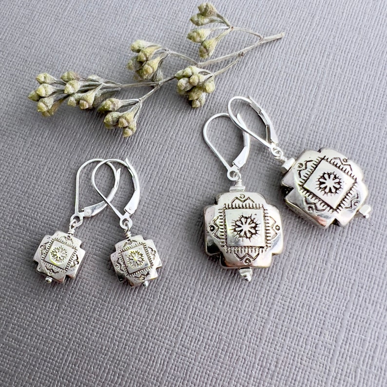 May include: Three pairs of silver dangle earrings with a square design featuring a detailed floral pattern. The earrings have a hook closure.