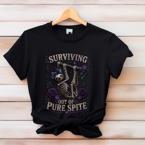 May include: Black t-shirt with a skeleton sloth design hanging from a branch, surrounded by purple roses and the text "SURVIVING OUT OF PURE SPITE". The sloth has a blue bow.