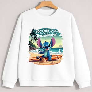 Puede incluir: Sudadera blanca con un personaje de dibujos animados en una playa. El personaje azul con orejas grandes est&aacute; sobre una tabla de surf. El texto "Too Cute to be this Troublemaker" est&aacute; encima del personaje.