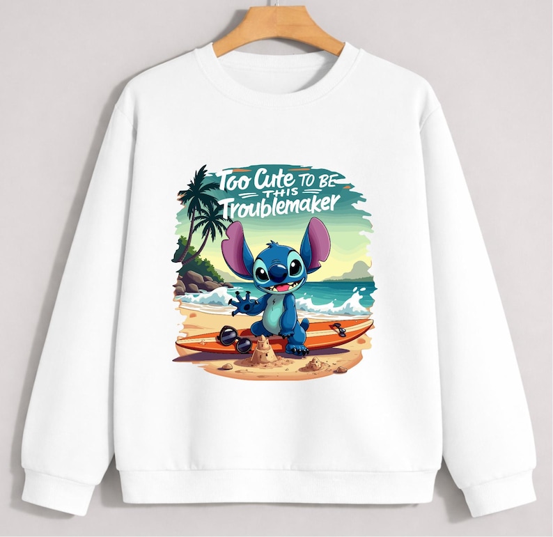 Puede incluir: Sudadera blanca con un personaje de dibujos animados en una playa. El personaje azul con orejas grandes est&aacute; sobre una tabla de surf. El texto "Too Cute to be this Troublemaker" est&aacute; encima del personaje.