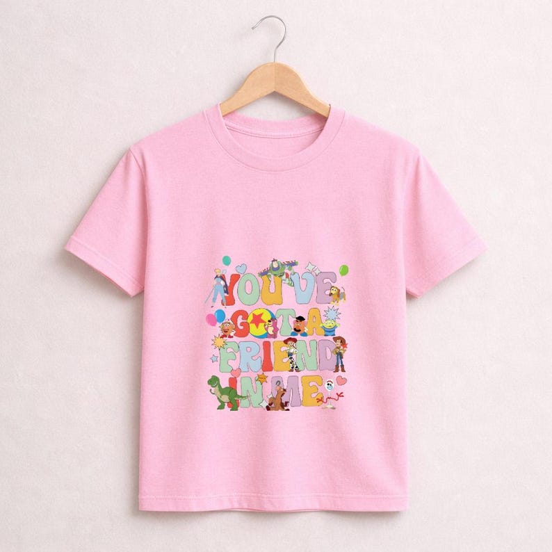 May include: A light pink t-shirt featuring the phrase "YOU'VE GOT A FRIEND IN ME" in colorful letters. The shirt showcases various cartoon characters, including Woody, Buzz Lightyear, and others, from the Toy Story franchise.